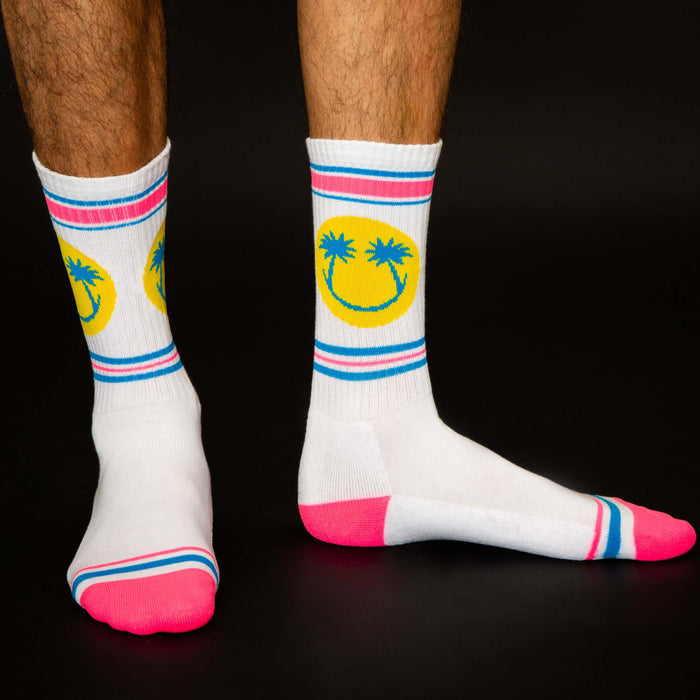 A pair of white socks with a colorful pattern of palm trees and smiley faces. The socks have pink toes and heels with blue and white stripes around the top.