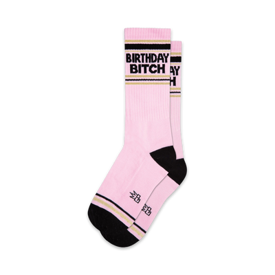 Two pink crew socks with black 'Birthday Bitch' lettering and gold stripes. Black and gold accents at the cuffs.