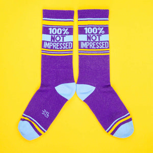 A pair of purple '100% Not Impressed' socks laid out in an X shape on a yellow background.