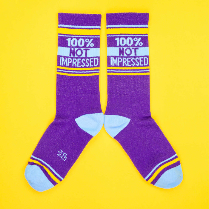A pair of purple '100% Not Impressed' socks laid out in an X shape on a yellow background.