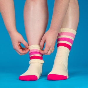 Lifestyle shot: A person putting on a pair of cream socks with pink and red stripes against a blue background.