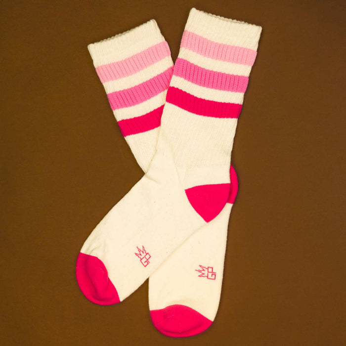 Flatlay product shot: Pair of cream crew socks with pink and red stripes on a brown background.