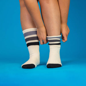 Lifestyle shot showing a person putting on a pair of off-white crew socks with black and grey stripes against a blue background.