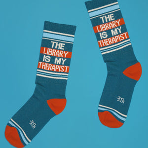 Pair of teal 'The Library Is My Therapist' socks on a blue background, angled view.