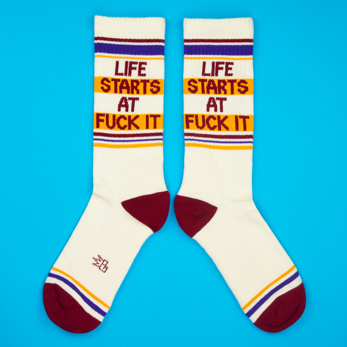 Flatlay product shot of a pair of 'Life Starts At Fuck It' socks on a bright blue background.