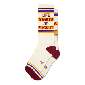 Pair of cream crew socks with maroon, gold, and purple stripes and the phrase 'Life Starts At Fuck It' in a retro font.