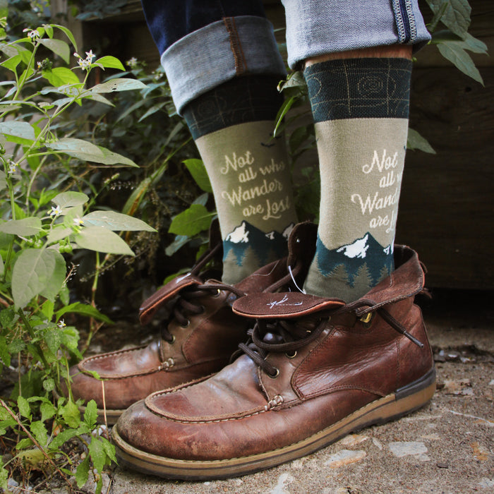 A person wearing brown leather boots, blue jeans, and green socks with the saying 