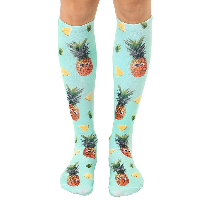 Pair of knee-high socks. Teal background with repeating pattern of cartoon pineapples with googly eyes and pineapple slices.