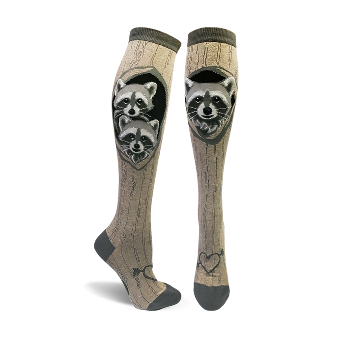 A pair of tan knee-high socks with a wood grain pattern and two raccoons peeking from a tree hole.  The socks have dark gray toes and heels, and a small heart with arrow detail at the ankle. }}
