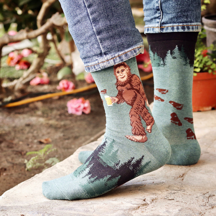 A person is wearing a pair of green socks with a pattern of bigfoot holding a beer on them. The socks are pulled up over the person's blue jeans.