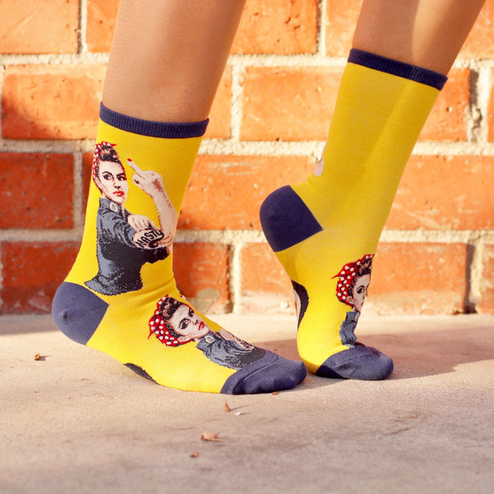 Lifestyle shot of a person wearing yellow 'Nasty Rosie' socks against a brick wall background.