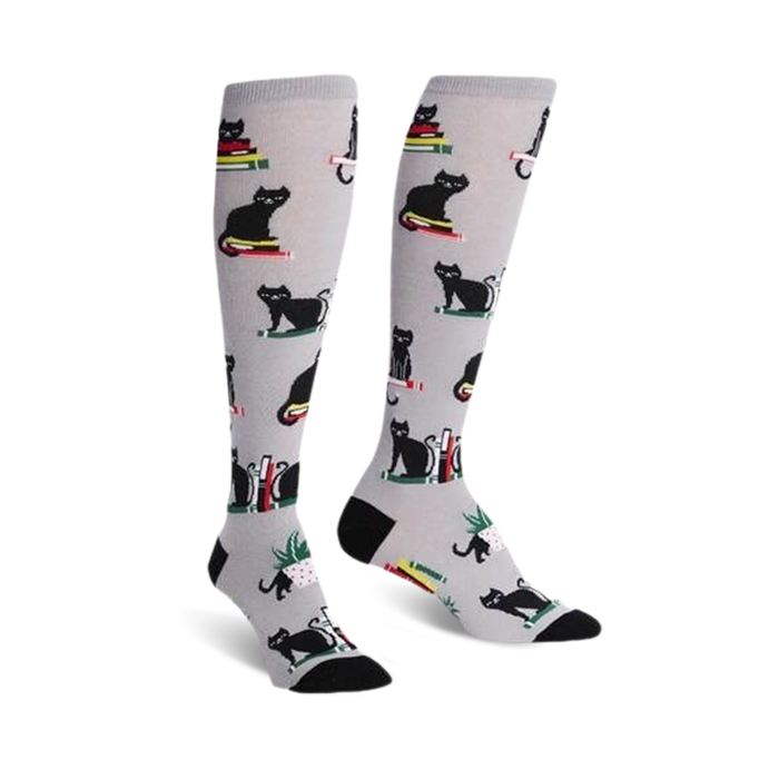 Pair of knee-high socks on a white background. They're light grey with a pattern of black cats sitting on stacks of colorful books. }}