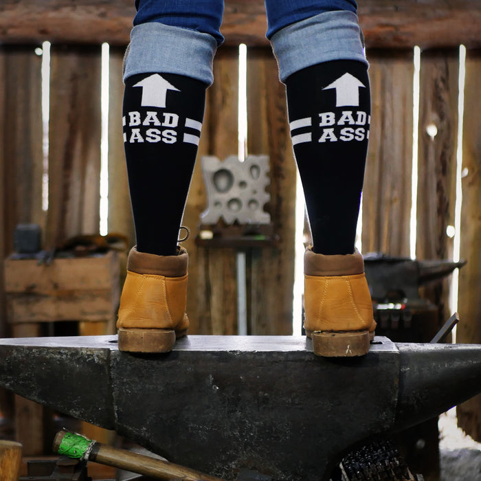 Lifestyle shot: Person wearing black 'Bad Ass' knee-high socks and work boots standing on an anvil.