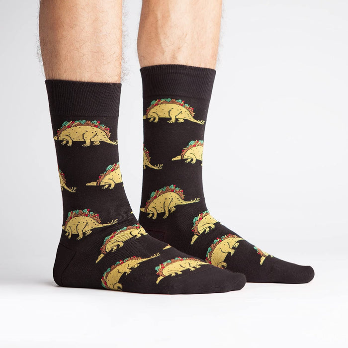 Lifestyle photo showing Tacosaurus socks on a person's feet. The socks are black with a fun taco-dinosaur pattern.