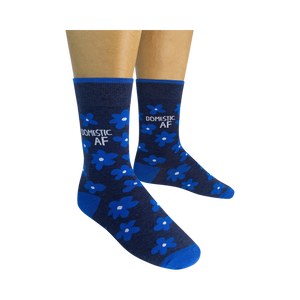 Pair of navy blue crew socks with bright blue flowers and 'DOMESTIC AF' text, shown on a model's feet.