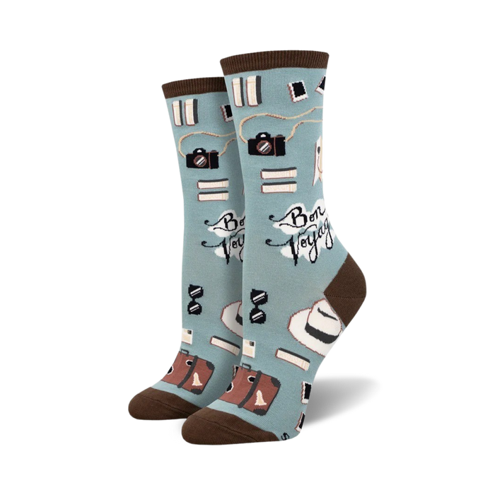A pair of crew socks in teal and mint green, featuring cameras, books, hats, and suitcases; the phrase 'Bon Voyage' is printed on the socks.