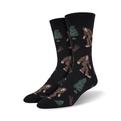 Pair of crew socks; black background with brown Bigfoot, green trees, and brown footprints. Fun, whimsical design.
