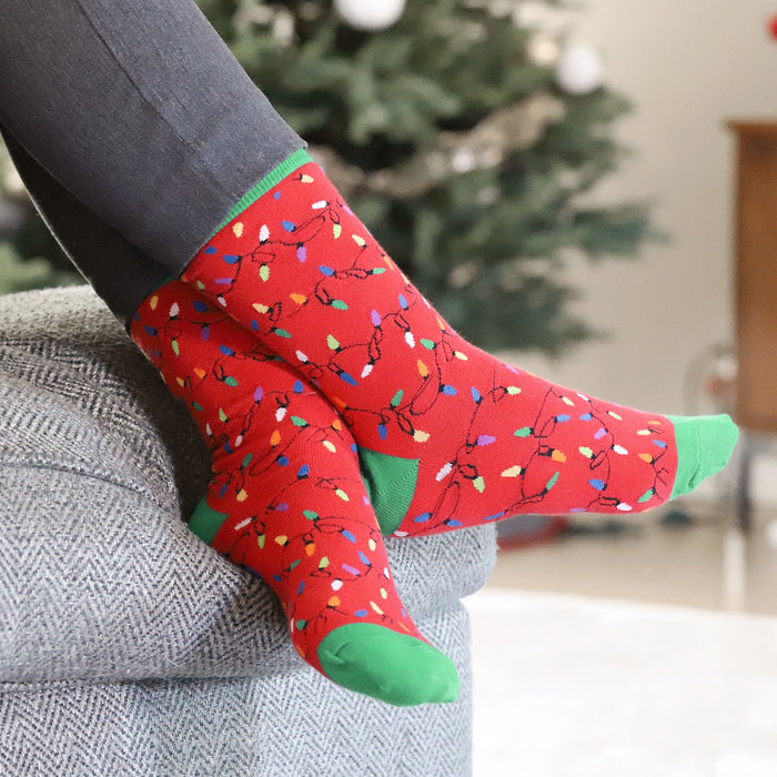 A pair of legs with red and green patterned socks on is resting on the floor in front of a decorated Christmas tree. A string of multicolored lights is wrapped around the legs. There are wrapped presents under the tree.