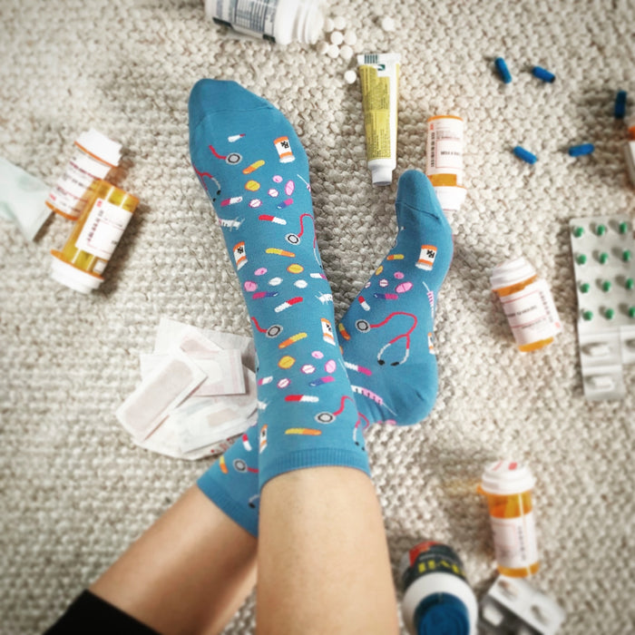 A person is wearing blue socks with a colorful pattern of pills, capsules, and syringes on them. The socks are resting on a white carpet, and there are various pill bottles, a tube of ointment, and some pills scattered on the carpet.