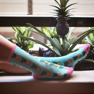 A person is sitting on a ledge with their feet resting on a concrete surface. They are wearing pink socks with a blue background and a pattern of yellow pineapples. The socks have a pink toe and heel. The person's legs are crossed at the ankles. In the background is a pineapple plant with green leaves.