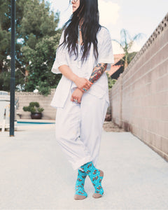 A young woman with long black hair and tattoos on her arms is crouching next to a swimming pool. She is wearing a white shirt, white pants, and colorful socks with otters on them. She has several rings on her fingers and a necklace around her neck.