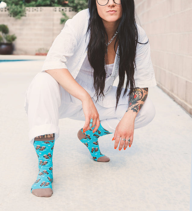 A young woman with long black hair and tattoos on her arms is crouching next to a swimming pool. She is wearing a white shirt, white pants, and colorful socks with otters on them. She has several rings on her fingers and a necklace around her neck.
