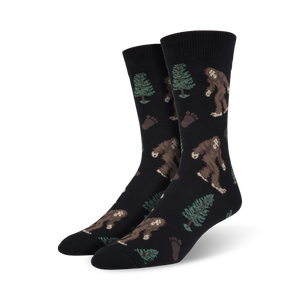 Pair of black Bigfoot XL crew socks. Features repeating pattern of Bigfoot, trees, and footprints. Brown, green, and black design on a black background.