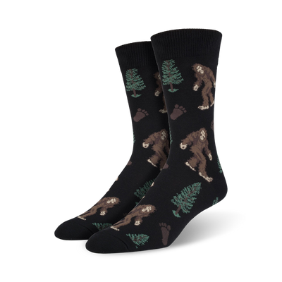 Pair of black Bigfoot XL crew socks. Features repeating pattern of Bigfoot, trees, and footprints. Brown, green, and black design on a black background.