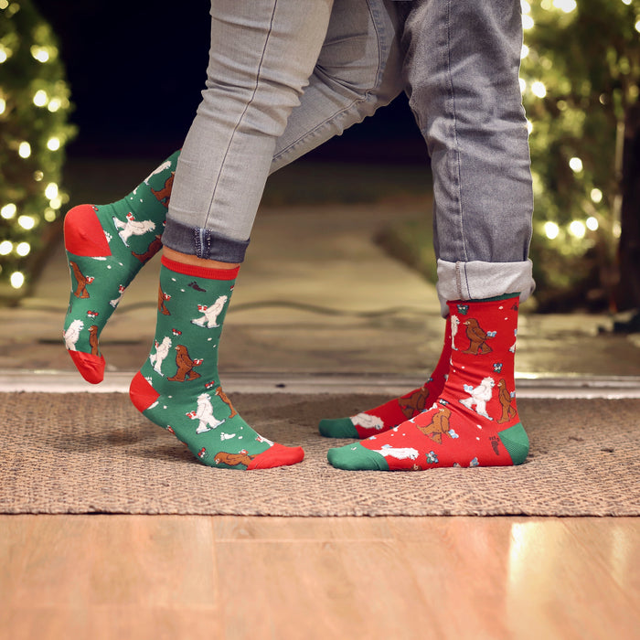 A man and a woman are standing on a brown patterned rug inside a house. The man and woman are wearing jeans and colorful socks with a yeti design. The background is blurry, but there are Christmas lights visible.