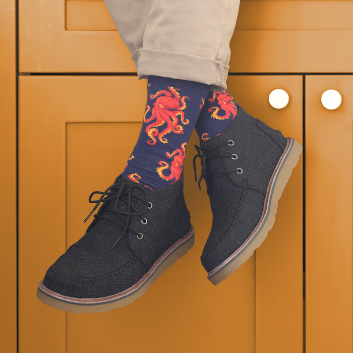 A person is wearing a pair of blue socks with a pattern of red octopuses and dark grey suede shoes. The person is sitting on an orange cabinet with their feet up. Only the person's legs, feet, and shoes are visible.