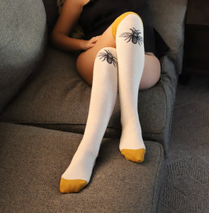 Lifestyle shot: A person wearing the off-white knee-high socks with mustard yellow cuffs and bee design, relaxing on a grey couch.