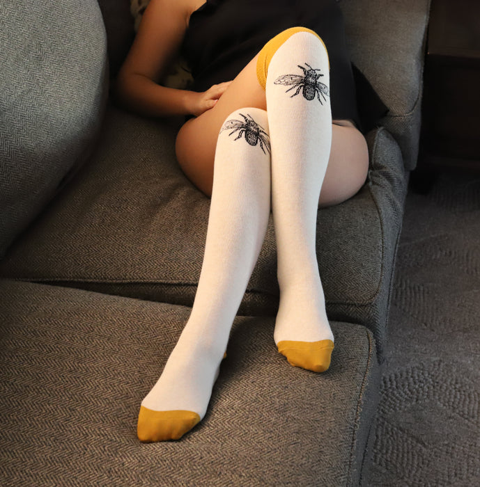 Lifestyle shot: A person wearing the off-white knee-high socks with mustard yellow cuffs and bee design, relaxing on a grey couch.