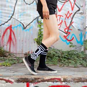 Lifestyle shot: Person wearing black '00 Fucks Given' socks with black Vans shoes against a graffitied wall.