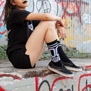 Lifestyle shot: Person wearing black '00 Fucks Given' socks and black shoes, standing against a graffitied wall.