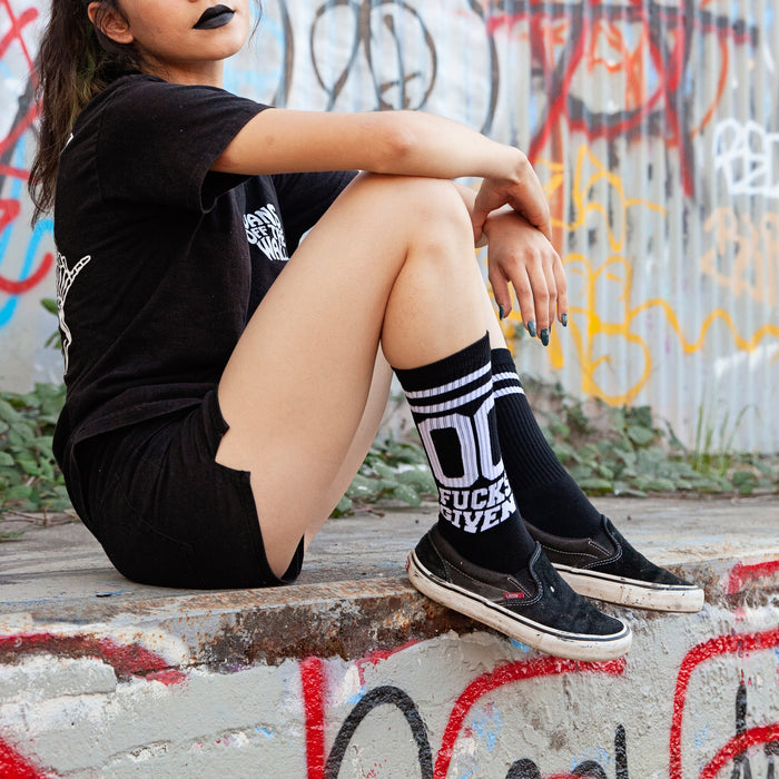 Lifestyle shot: Person wearing black '00 Fucks Given' socks and black shoes, standing against a graffitied wall.