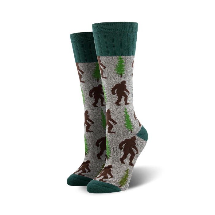A pair of grey crew socks with brown Bigfoot and green trees. The socks have teal cuffs. }}
