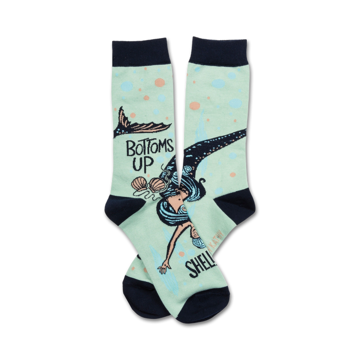 A pair of mint green crew socks featuring a whimsical illustration of a mermaid swimming upside down with seashells. The socks also say 'Bottoms Up'. }}