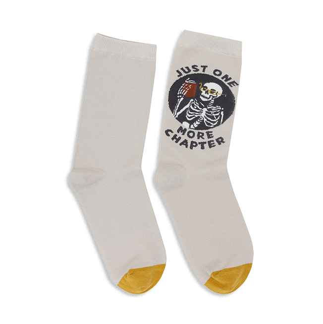 Pair of off-white crew socks with a mustard yellow heel and toe. A circular design shows a skeleton reading a book with the text 'Just One More Chapter'. }}