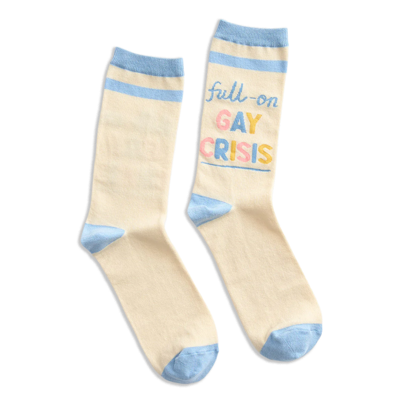 A pair of cream-colored crew socks with light blue accents on the heels, toes, and cuffs, featuring the text 'full-on GAY CRISIS' printed on one sock in pastel blue, pink, and yellow lettering.