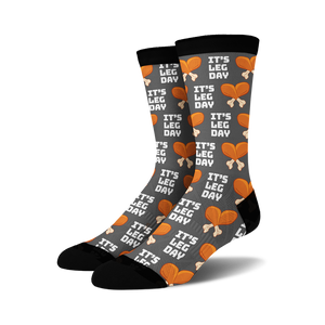 Pair of crew socks, dark grey with orange and white 'It's Leg Day' text and chicken leg illustrations repeating across the fabric.
