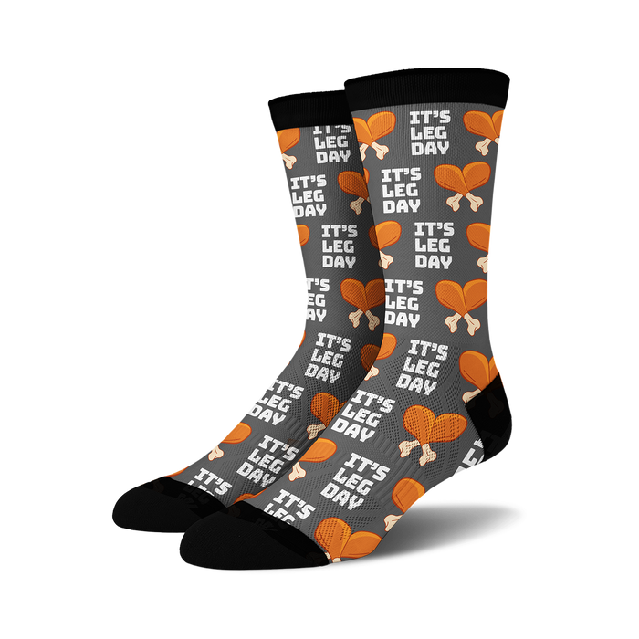 Pair of crew socks, dark grey with orange and white 'It's Leg Day' text and chicken leg illustrations repeating across the fabric.