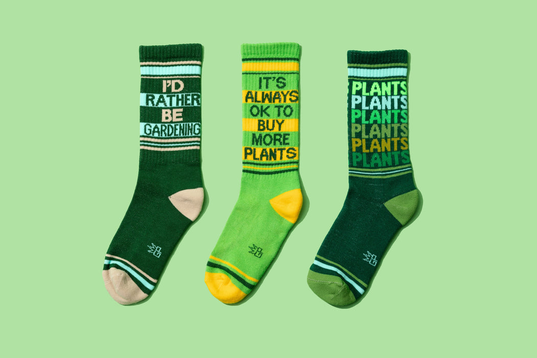 Product shot of three different gardening-themed socks: 'I'd Rather Be Gardening', 'It's Always Ok To Buy More Plants', and 'Plants Plants Plants'.