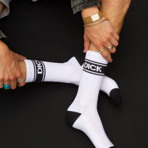 Lifestyle shot: A person seated with legs crossed, wearing 'Dick' socks and silver Converse sneakers. Dark background.