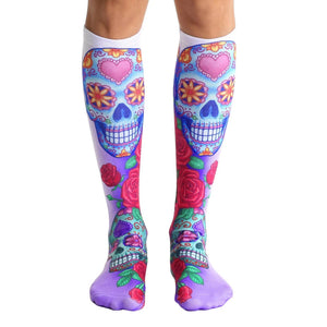 Pair of knee-high socks.  The main design features colorful sugar skulls with pink hearts and roses on a white and purple background.