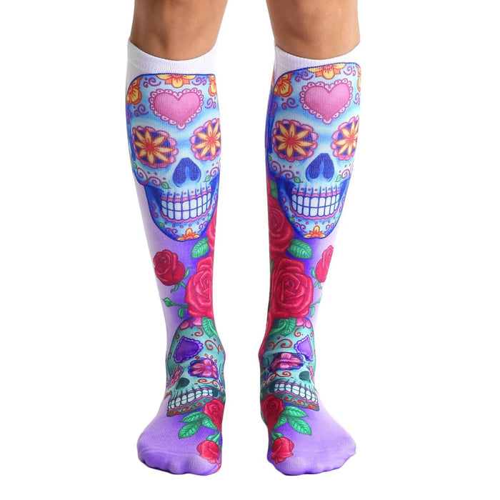 Pair of knee-high socks.  The main design features colorful sugar skulls with pink hearts and roses on a white and purple background.