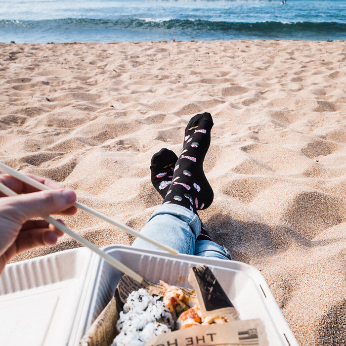 A person is sitting on the beach with their feet in the sand. They are wearing jeans and sushi socks and holding chopsticks. There is a container of sushi on the sand in front of them. The ocean is in the background.