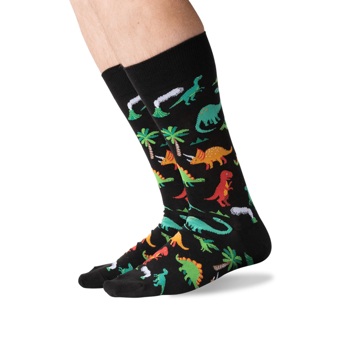 A pair of black socks with a pattern of colorful dinosaurs, palm trees, and volcanoes.