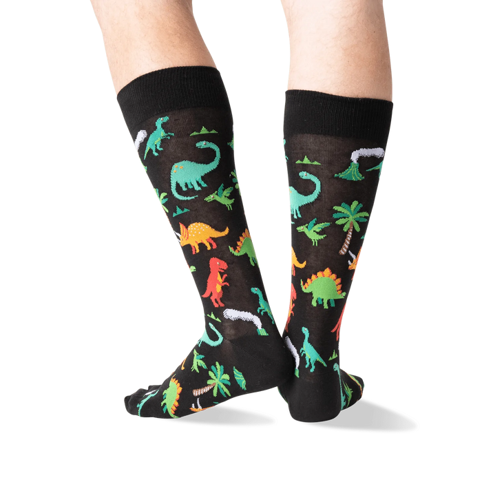 A pair of black socks with a pattern of colorful dinosaurs, palm trees, and volcanoes.