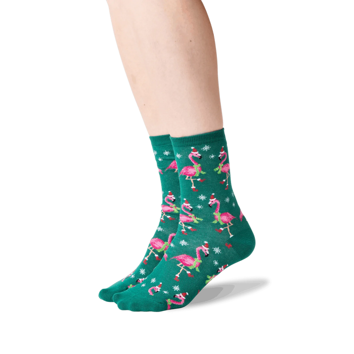 A pair of green socks with a pattern of pink flamingos wearing Santa hats and scarves. The socks are shown on a person's legs.