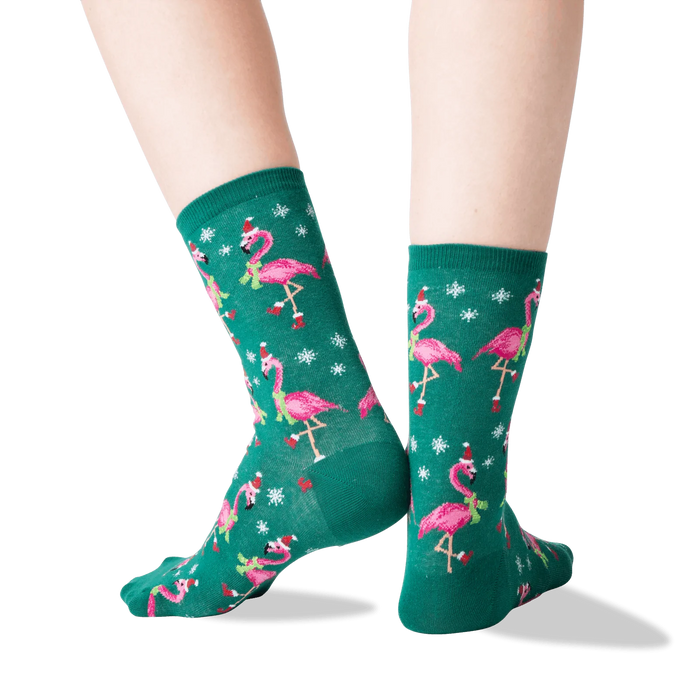 A pair of green socks with a pattern of pink flamingos wearing Santa hats and scarves. The socks are shown on a person's legs.